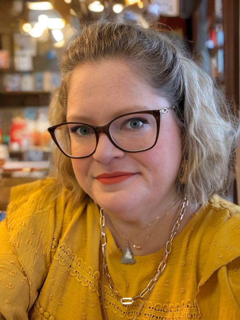 Brande Wester, smiling woman with glasses and wavy hair, wearing a yellow top and layered necklaces.